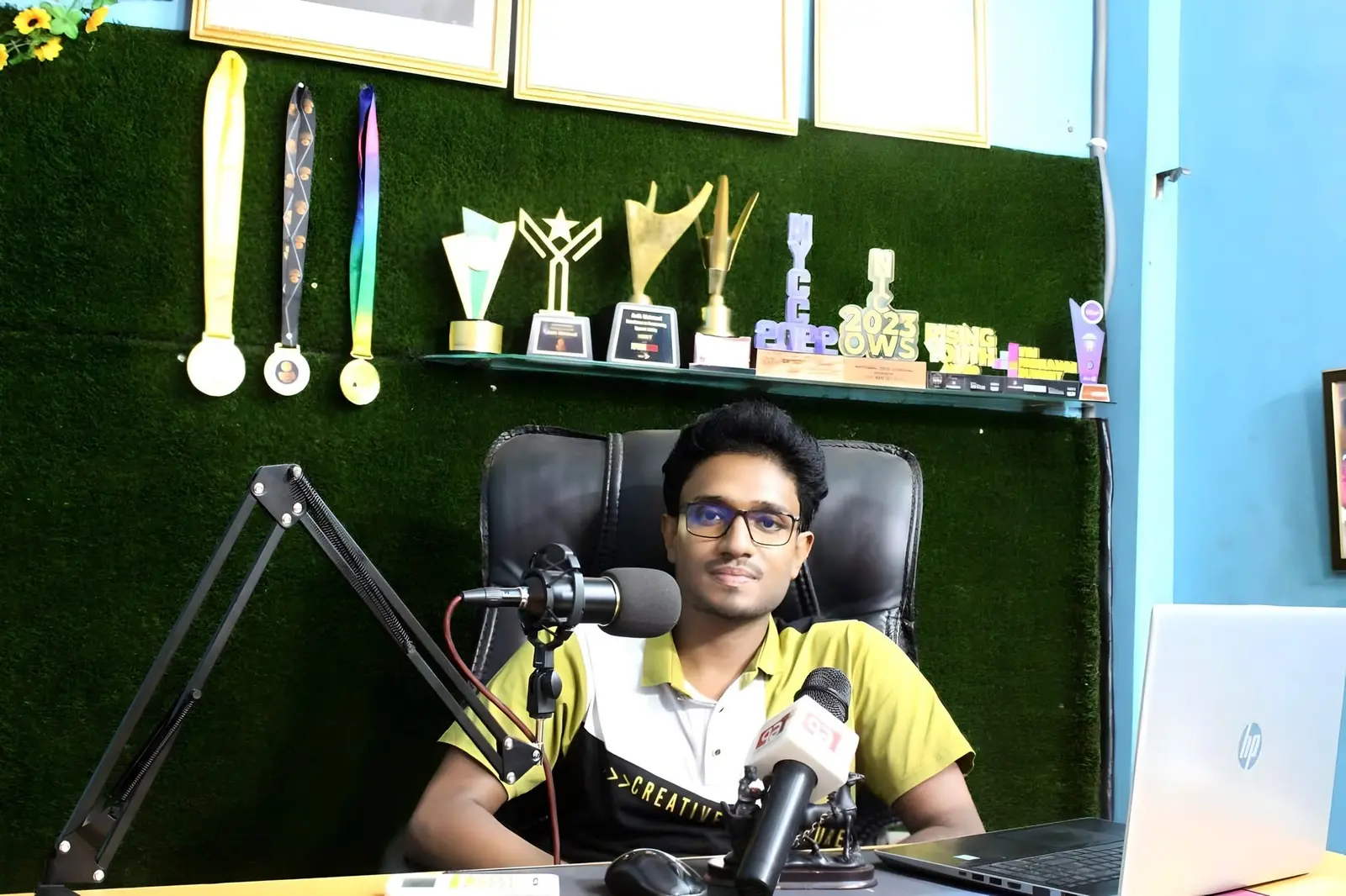Anik Mahmud speaking into studio and media microphones at his Professor IT Zone workspace, with awards displayed behind him.
